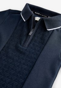 Marineblauw poloshirt met gestructureerd geometrisch patroon aan de voorkant, halve ritssluiting met "Baker" trekker, en een kraag met witte rand.
