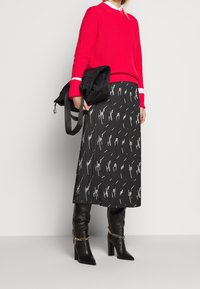 Mujer con suéter rojo, falda midi negra con texto en blanco, botas de tacón negras con cadenas doradas, sosteniendo un bolso negro plegado.
