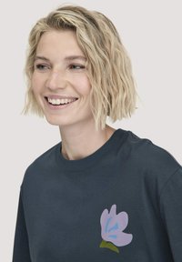 Glimlachende vrouw met kort golvend blond haar, gekleed in een donkerblauwe shirt met een kleine paarse en blauwe bloemenafbeelding op de borst.