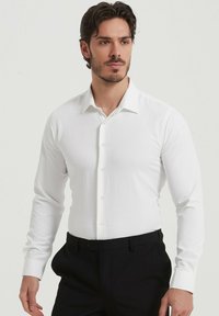 Chemise blanche à manches longues avec un col classique et des boutons, fabriquée en tissu lisse. Portée avec un pantalon noir à coupe ajustée.
