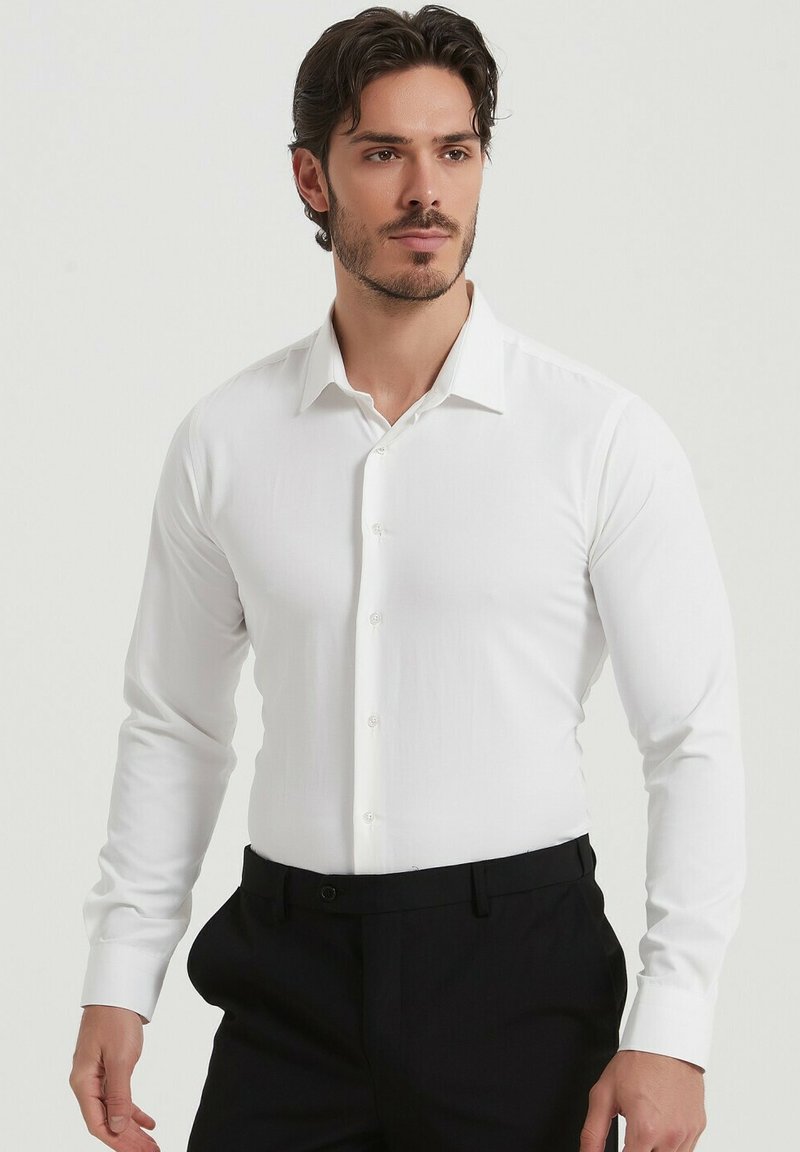 Chemise blanche à manches longues avec un col classique et des boutons, fabriquée en tissu lisse. Portée avec un pantalon noir à coupe ajustée.