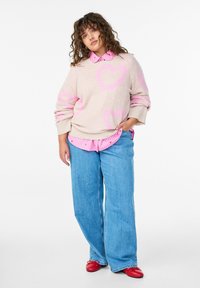 Lokkis juustega naine, kes kannab beežit kampsunit roosade südametega, roosade triibulise särgiga, laia siniste teksade ja punaste kingadega valgel taustal.