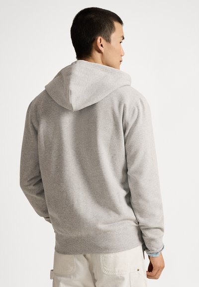 Άνδρας που φοράει ανοιχτό γκρι φούτερ με κουκούλα και άσπρο παντελόνι, στραμμένος προς τα πίσω με φόντο απλό λευκό.