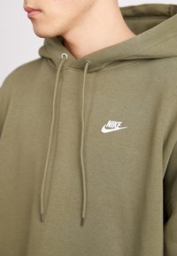 Olīvu zaļš kapučjaka krekls ar aizsietām auklām, izgatavots no mīksta kokvilnas. Tam ir balts Nike logo uz krūšu daļas un brīvā fit dizains.