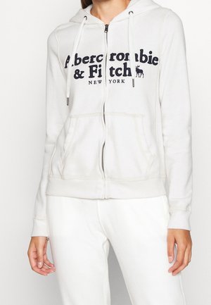 Haut de survêtement zippé beige clair avec poches avant, capuche à cordon et impression du logo navy. Le tissu est doux et lisse, avec des poignets et un ourlet côtelés.