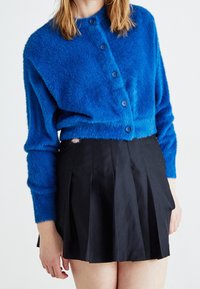 Niebieski, puszysty kardigan z okrągłym kołnierzem i sześcioma guzikami, w połączeniu z plisowaną czarną spódniczką mini z małą metką z logo.
