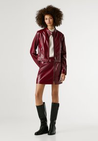 Bordeaux faux leren jas met een rits aan de voorkant, gestyled over een bijpassende pencil skirt, gecombineerd met zwarte kniehoge laarzen. Glad, glanzend textuur.