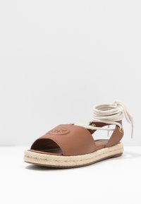 Sandale espadrille en cuir marron avec bout ouvert et liens à la cheville. Elle possède une semelle en corde de jute et un logo en relief sur le dessus.