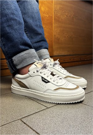 Baskets en cuir blanc avec des accents beiges portées avec un jean bleu retroussé sur un sol carrelé à l'intérieur.