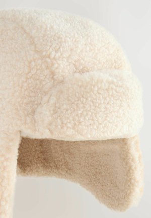 Krämfärgad fuskpälsmössa med öronlappar, som har en mjuk, texturerad yta och en rundad toppdesign för värme och skydd.