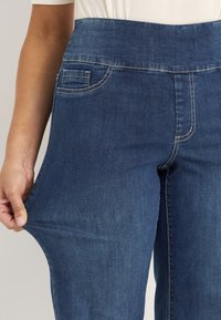 Persoon in blauwe stretch denim jeans die de stof naar buiten trekt om de flexibiliteit en pasvorm te tonen, met zichtbare witte stikdetails.