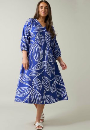 Sininen mekko valkoisilla lehtikuvioilla, V-kaula-aukko ja kyynärpituiseen hihaan. Virtaviivainen A-linja, jonka kangas on teksturoitu, sopii arkisiin asuihin.