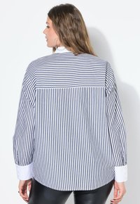 Camicia a righe con bottoni, caratterizzata da righe verticali blu navy e bianche, maniche lunghe e polsini bianchi. Tessuto liscio con una vestibilità rilassata.