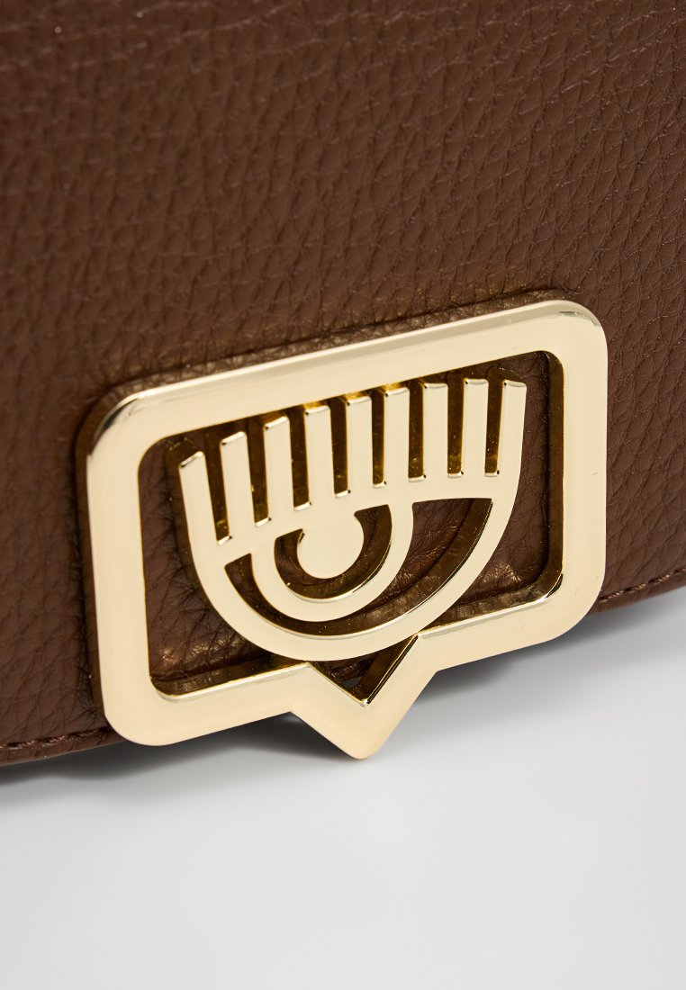 Bolso de cuero marrón con textura, que presenta un prominente emblema en forma de ojo de metal dorado, con líneas horizontales y una base triangular.