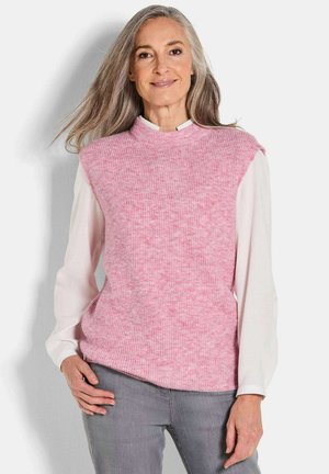 Vrouw met lang grijs haar, gekleed in een roze mouwloze gebreide vest over een witte blouse en grijze broek, staand tegen een effen achtergrond.