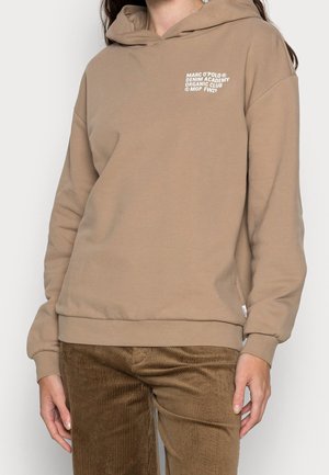 Person wearing a light brown hoodie with white text on the chest and brown corduroy pants, standing with arms relaxed by sides.