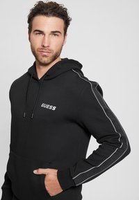 Black hoodie made of cotton, featuring a front pocket, drawstrings, and white side stripes. Embroidered "GUESS" logo on chest.