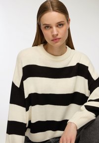 Sweter w paski w kolorze czarnym i kremowym, o luźnej, dzianinowej strukturze. Posiada rypsowy dekolt typu crew neck oraz szerokie rękawy dla swobodnego fasonu.