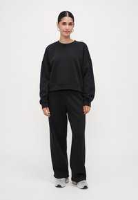 Donna con capelli scuri, in una felpa nera e pantaloni a gamba larga abbinati, con sneaker grigie chiare, in piedi davanti a uno sfondo semplice.