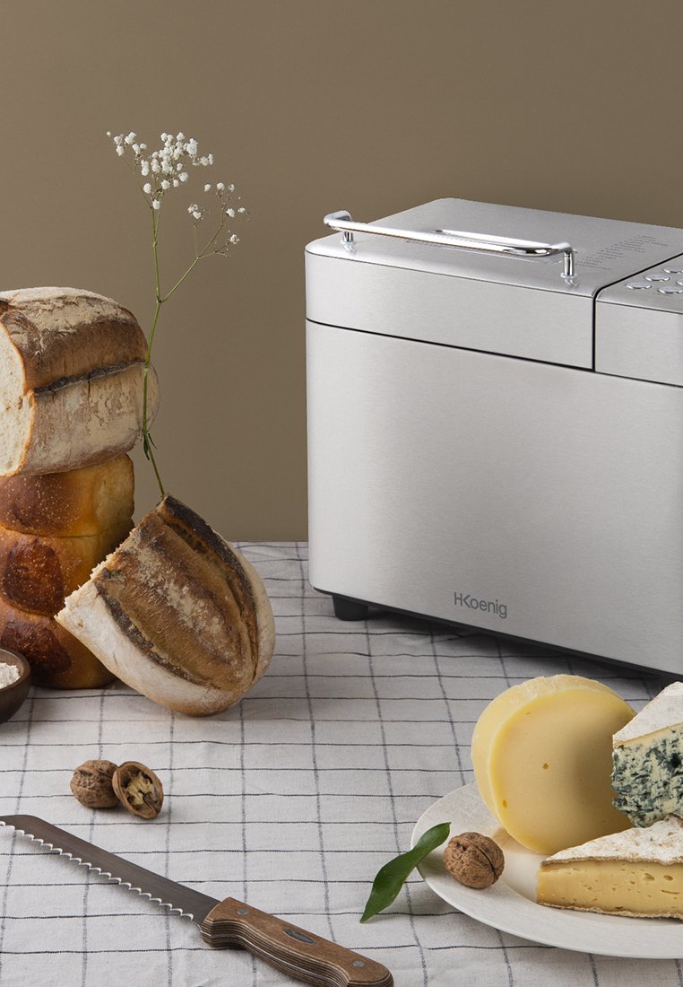 Roestvrijstalen broodrooster naast verschillende broden en kazen. Inclusief een zaagmes en een houten kom met noten op een geruite doek.