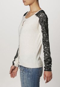 Kvinna iklädd en vit cardigan med svarta blommiga spetsärmar, i kombination med blå jeans, stående i profil mot en neutral bakgrund.