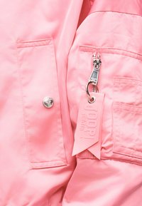 Κοντινή λήψη υφάσματος ροζ μπουφάν που δείχνει φερμουάρ με μεταλλική ταινία έλξης και προσαρτημένη ροζ ετικέτα από ύφασμα, ένα κουμπί τύπου snap και ραφές στολίδι στην τσέπη.