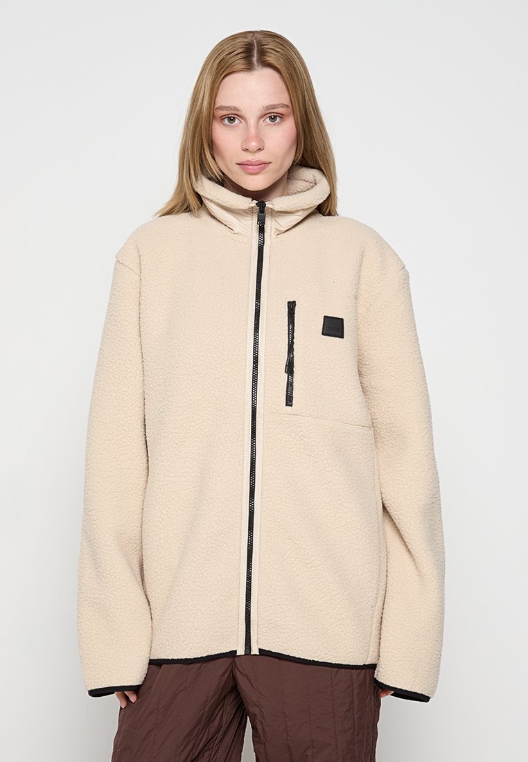 Beige Fleecejacke mit hohem Kragen, Frontreißverschluss und einer mit Reißverschluss versehenen Brusttasche. Verfügt über schwarze Akzente am Reißverschluss und am Saum.