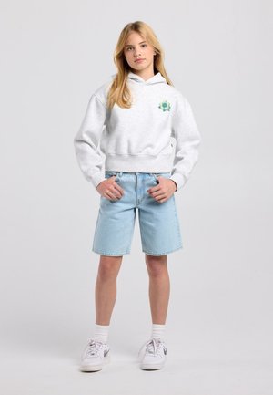 Fille portant un sweat à capuche gris avec un emblème de fleur verte, un short en jean bleu clair, des chaussettes blanches et des baskets blanches, debout les mains sur les hanches.