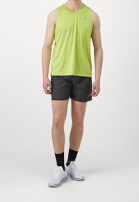 Zelený bezrukávový sportovní top, černé šortky, bílé boty s šedými akcenty a černé ponožky. Hladká tkanina, neformální design a přiléhavý střih.