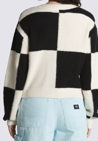 Pletený svetr s černo-krémovým kostkovaným vzorem, s volným střihem a žebrovanými rukávy, vrstvený přes světle modré pracovní kalhoty.