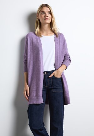 Cardigan lilas en tissu doux avec des poignets et un ourlet côtelés, porté sur une chemise blanche et associé à un jean bleu foncé.