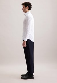 Un uomo sta di profilo indossando una camicia bianca aderente, pantaloni scuri su misura e scarpe eleganti nere, su uno sfondo chiaro e semplice.