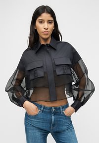 Γυναίκα που φοράει διάφανη μαύρη κοντή μπλούζα με φουσκωτά μανίκια και πλαϊνές τσέπες με καπάκι, συνδυασμένη με μπλε τζιν, πόζα με τα χέρια στις τσέπες.