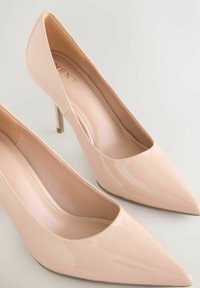 Naakte lakleren pumps met hoge hak en puntige neus, met een gladde textuur en een naaldhak. Zachte binnenvoering zichtbaar.