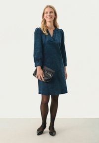 Navyblaues Cordkleid mit V-Ausschnitt, Puffärmeln und einem Saum oberhalb des Knies. Kombiniert mit einer schwarzen Clutch und schwarzen Strumpfhosen.