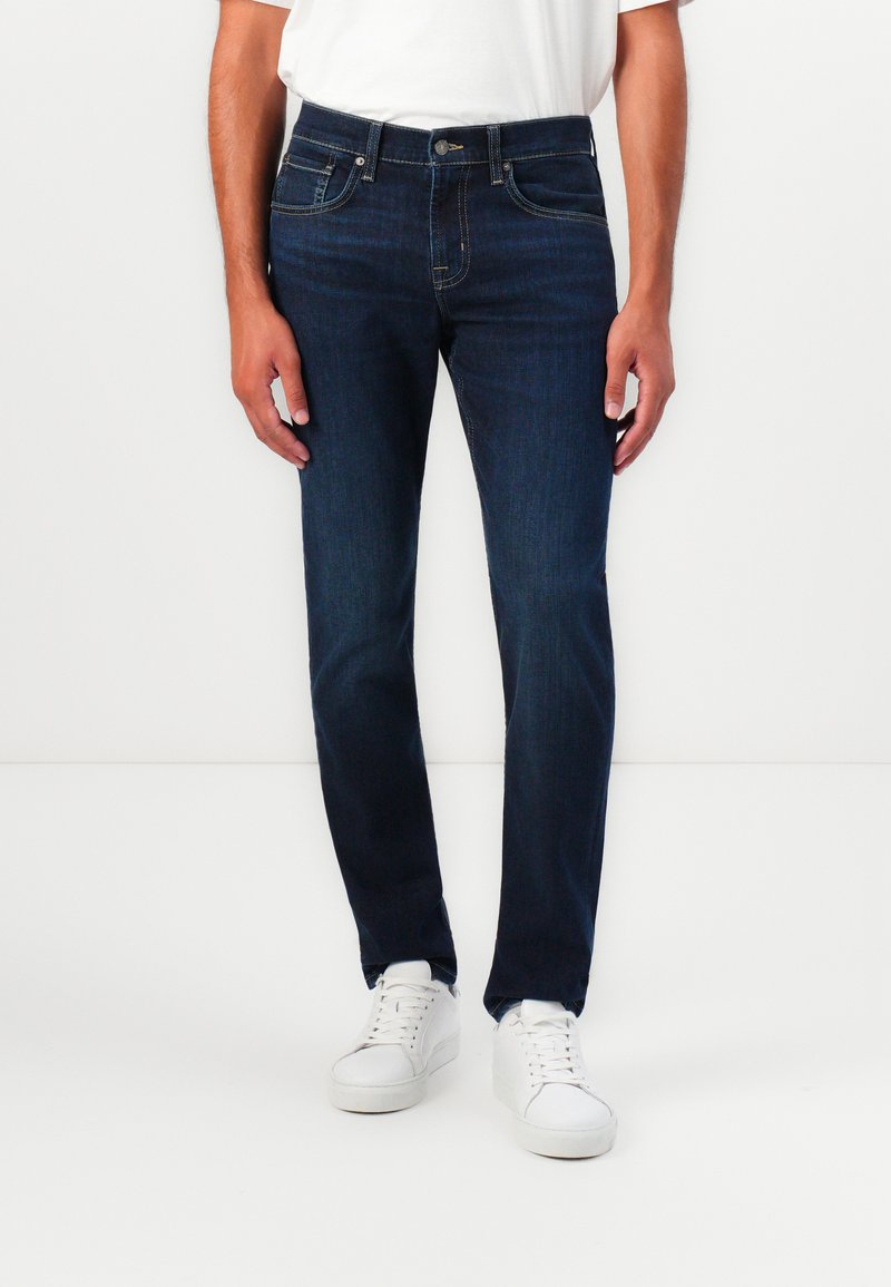 Jeans in denim blu scuro con taglio dritto, dotati di chiusura con bottoni e zip, cinque tasche e dettagli di cucitura sottili. Accoppiati con scarpe da ginnastica bianche.