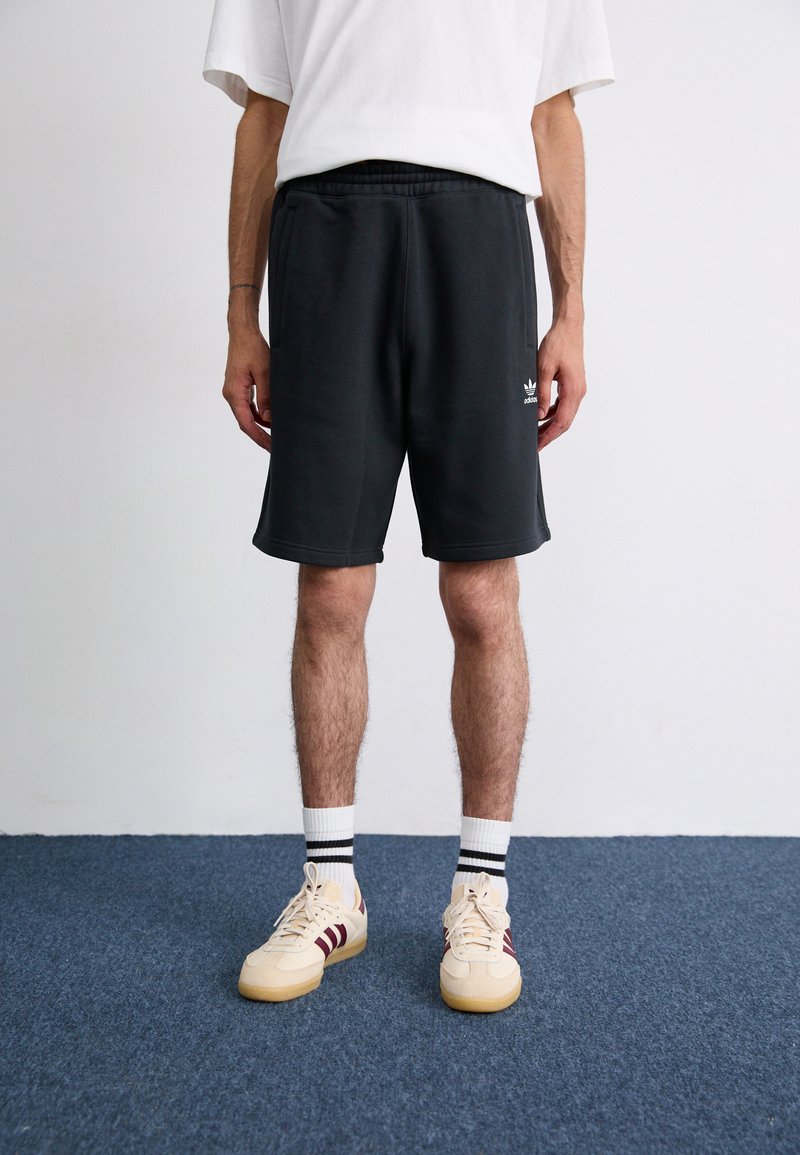 Black shorts with an elastic waistband, featuring side pockets and a small white logo. Worn with beige sneakers and striped socks.