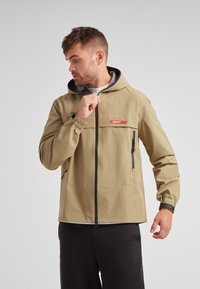 Chaqueta con capucha de un tono marrón claro, con cremallera en el frontal, puños elásticos y un bolsillo en el pecho que presenta una etiqueta roja. Tejido suave y resistente al agua.