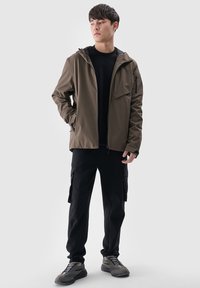 Jeune homme debout portant une veste marron à capuche, un t-shirt noir, un pantalon cargo noir et des baskets grises, regardant légèrement sur le côté.