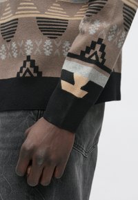 Temnopolta roka ob bež, črni in sivi vzorčasti rokav puloverja ter izpranih črnih kavbojkah iz jeansa.