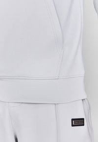 Lichtgrijze sweatshirt met een zachte textuur, voorzien van een hoge hals, geribbelde manchetten en een klein zwart merklabel aan de zijkant.