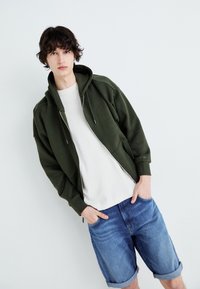 Chaqueta con capucha y cremallera en verde oscuro, con una textura suave y cordones ajustables. Combinada con pantalones cortos de mezclilla claros y una camisa color crema.
