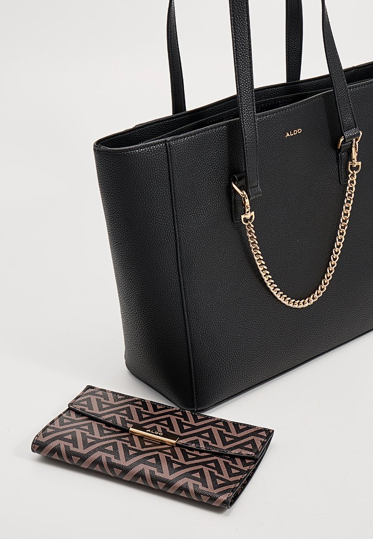 Bolso tote negro con textura y acento de cadena, equipado con dos asas resistentes, acompañado de una cartera con patrón geométrico en tonos marrones y negros.