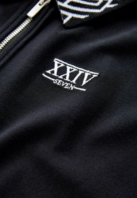 Sort polo skjorte med lynlås, som har et struktureret stof, et mønstret krave og hvidt broderet logo "XXIV SEVEN."
