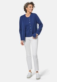 Blauer Cardigan mit Knopfverschluss, getragen über einem blauen Oberteil, kombiniert mit weißen Hosen und weißen Sneakers. Lockige Haare, im Stehen.