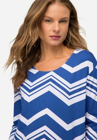 Niebieska i biała bluzka z długim rękawem, z wzorem w zygzak, okrągłym dekoltem i miękką teksturą. Projekt podkreśla kontrastujące kolory i kształty.
