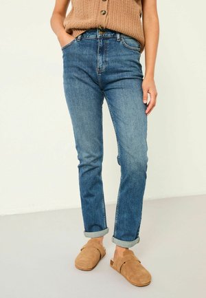 Person trägt hochgekrempelte blaue Jeans, eine braune ärmellose Strickjacke und beige Wildleder-Slipper, steht mit einer Hand in der Tasche.