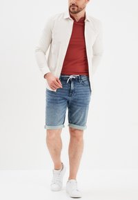 Camisa de manga larga a rayas blancas sobre una camiseta roja, combinada con pantalones cortos de mezclilla azul con dobladillos enrollados y zapatillas blancas.