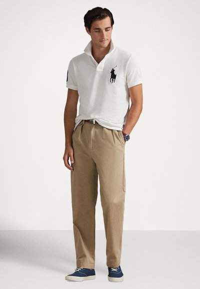 Polo branco com logótipo preto, calças cáqui plissadas e sapatos de lona azuis. Tecido macio, ajuste casual, com mangas curtas e gola.