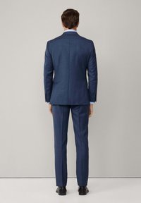 Marineblaues maßgeschneidertes Anzug mit glatter Textur, ausgestattet mit einem Schlitz hinten, kombiniert mit einem hellblauen Hemd und schwarzen Schuhen.